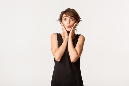 Close-up of surprised and amazed girl looking at camera impressed, standing in black dress over white background.