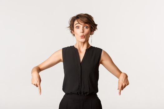Amazed attractive young woman pointing fingers down, gasping impressed, white background.