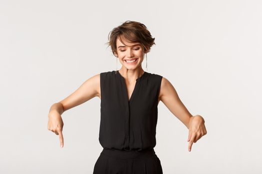 Happy adult woman in black dungarees pointing fingers down, looking downwards with pleased smile.