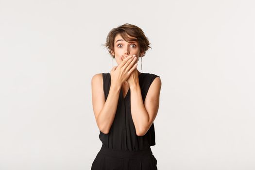 Shocked young woman found out secret, gasping and shut mouth with hands amazed, standing white background.