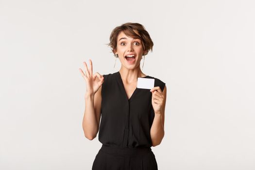Impressed attractive businesswoman showing okay gesture and credit card, standing over white background.