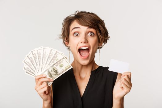 Close-up of amazed young woman open mouth fascinated, holding money and credit card, standing over white background.