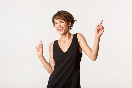 Image of carefree stylish woman having fun, dancing and singing, standing over white background.