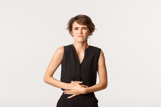 Woman in formal outfit touching belly and grimacing from pain. Girl having painful cramps or stomach ache, standing white background feeling discomfort.