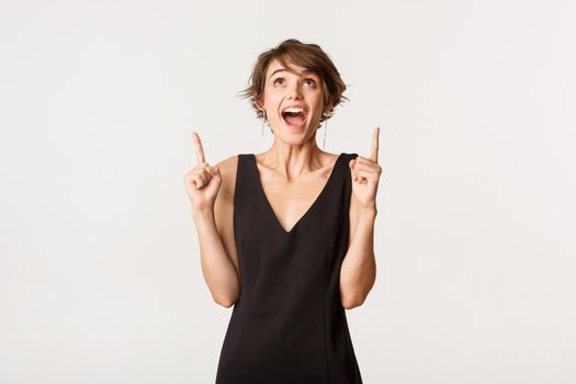 Excited attractive woman looking and pointing up with amazed ecstatic expression, standing over white background.