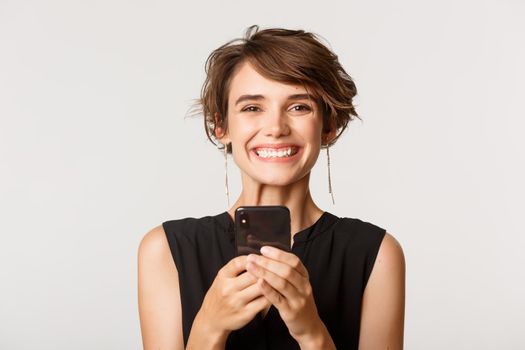 Close-up of happy woman holding smartphone, smiling at camera, standing over white background.
