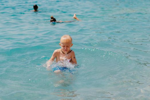 Child learning to swim in the open sea of tropical resort. Kids learn swimming. Exercise and training for young children. Little boy with colorful float board in sport club. Swimming baby or toddler. Happy child boy swims in sea in swimming circle with splash. Blue sky and water. Swimming training. Fun joy activities on vacation in the beach. Childhood moments lifestyle. Freedom careless. boy swim in the sea.