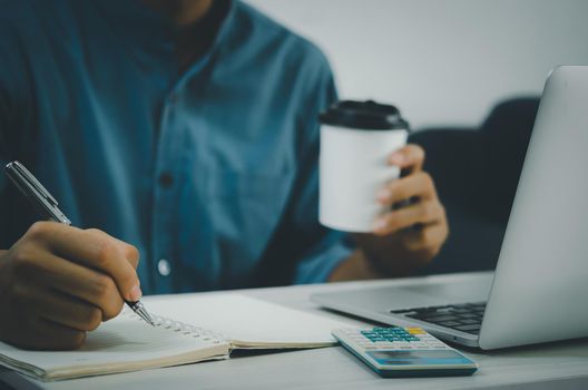 Male hand holding pen and writing at notebook and coffee cup on desk with notebook computer and calculator.