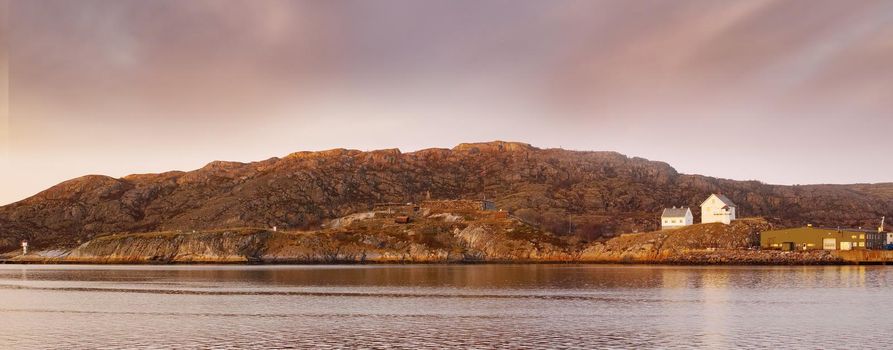 Sea landscape near a mountain with houses on a hill. The ocean or a large lake with residential buildings in the distance outdoors during sunset on a cloudy day. Perfect location for summer vacation.