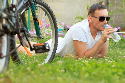 Close up view of man lying on green grass and drinking water from a bottle. Picture of male in white T-shirt and sunglasses with bottle of water lying on green grass with road bicycle beside him in the garden. Selective focus on man. Healthy active lifestyle