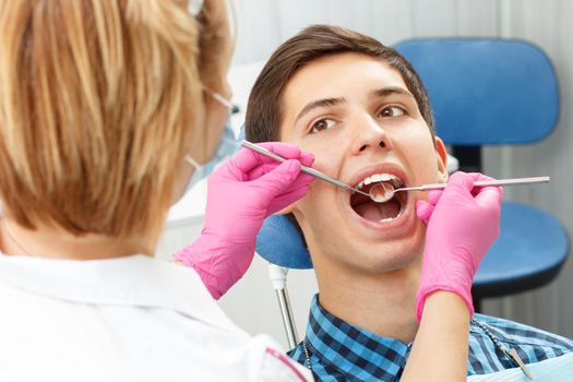 Handsome young man in visit at the dental office. He is sitting on the chair and dentist is examining his teeth with dental tools. Focus on patient mouth. Dentistry