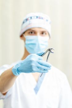 Portrait of handsome male dentist with stainless steel dental tongs or pliers for extraction of lower teeth in the dental clinic. Selective focus on dental tongs