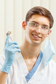 Portrait of handsome male dentist with stainless steel dental tongs or pliers and extracted lower tooth in it. Doctor wearing white uniform, glasses and blue gloves