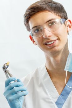 Portrait of handsome male dentist with stainless steel dental tongs or pliers and extracted lower tooth in it. Doctor wearing white uniform, glasses and blue gloves