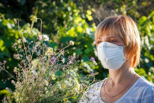 Unhappy woman in protection mask holding bouquet of wildflowers and trying to fight allergies to pollen. Woman protecting her nose from allergens. Allergy concept.