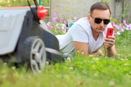 Close up view of man lying on green grass before mowing it. Picture of male in white T-shirt and sunglasses with mobile phone lying on grass with lawn mower beside him in the garden. Selective focus on man