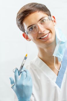 Portrait of handsome male dentist with syringe with anesthetic and needle in the dental clinic. Doctor wearing white uniform, glasses and blue gloves