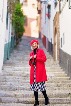 Full length of content positive young female traveler, in stylish outfit with photo camera on neck smiling brightly and looking at camera while standing near ages stone staircase in town