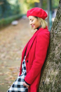 Side view of positive charming female in red coat and beret leaning on tree trunk in autumn park and looking down