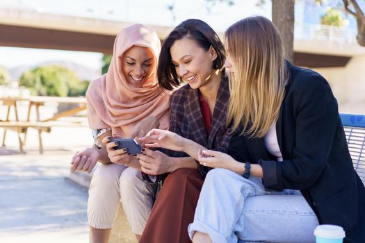 Content young multiracial women best friends, in casual clothes and hijab, smiling while watching video on smartphone sitting on bench in city park on sunny day