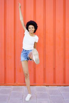 Full body of cheerful African American female with Afro hairstyle in casual wear looking at camera with raised leg on red background