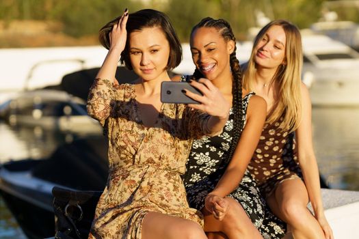 Group of delighted multiracial female friends taking selfie on smartphone while sitting together in port with boats on sunny summer day