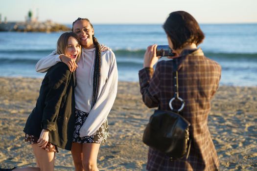 Anonymous female photographer with smartphone taking photo of cheerful multiracial female friends while standing on sandy beach near waving sea