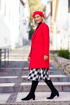 Full body of positive female in red coat and beret looking at camera while walking on stairs near buildings in city