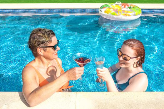 two women friends, toasting and drinking a cocktail by the pool of a resort on a sunny day. mother and daughter enjoying a summer holiday together. concept of holidays and travel. outdoor garden, natural sunlight.