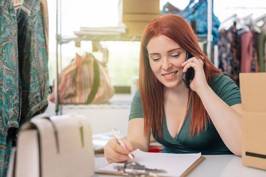 young businesswoman smiling, talking on her smartphone to her customers and taking down orders in the office of her clothing shop. work and business concept. natural light from window, background with clothes racks and clothes.