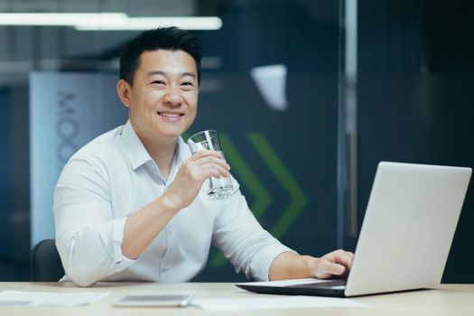 Asian businessman in the office drinking clean filtered water, portrait of a man smiling and looking at the camera holding a glass of water, a man at work with a laptop