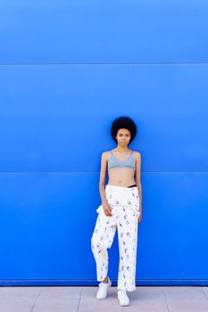 Full body of fit African American female in bra and overall looking at camera while standing against blue background on street