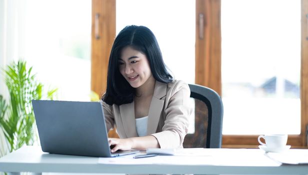 Happy young Asian businesswoman working on laptop keyboard with document at office .