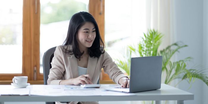 Asian businesswoman working in financial accounting analyzes a chart using a laptop calculator with documents and holding a pen sitting on a chair at an office desk.