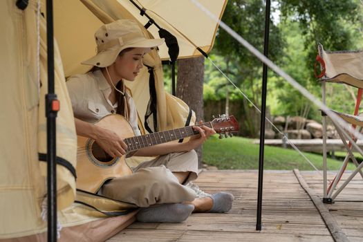 Tourist asian girl in hat sits in tent and playing guitar concept. Tourism rest on nature. Camping lifestyle.