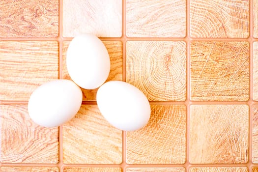 an oval or round object laid by a female bird, reptile, fish, or invertebrate, usually containing a developing embryo. Three chicken bird eggs on a wooden pink orange checkered surface.