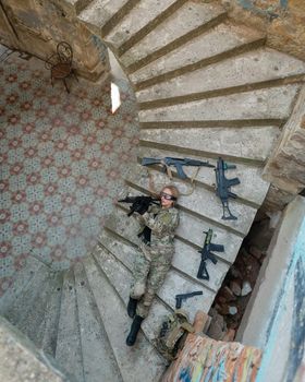 Caucasian woman in military uniform lies on the stairs of an abandoned building and holds a machine gun. View from above