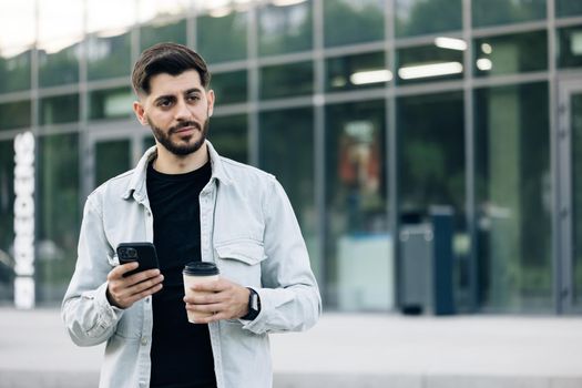 Bearded handsome arabian man having slow walk in the town, enjoying his tasty coffee. Typing messages on his smartphone and drinking hot coffee. Wearing stylish clothes. Having nice hairstyle.