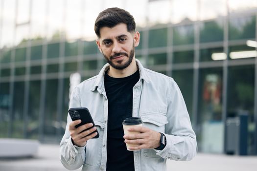 Attractive caucasian armenian young bearded man walks down the street uses phone smile sunlight sunset cellphone fashion internet face outside technology city mobile summer happy.