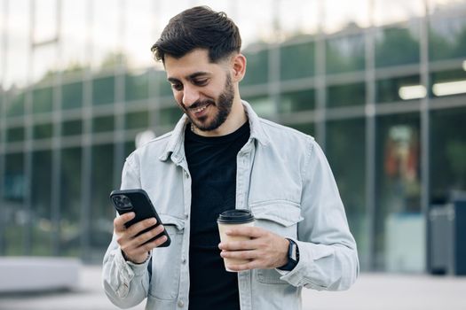 Young elegant bearded man using social media application on smartphone text messages receive news smiling outdoor. People portraits. Technology.