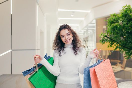 Portrait of a young curly beautiful woman who goes shopping in the mall and buys clothes. He holds colored paper bags in his hands, looks at the camera, smiles