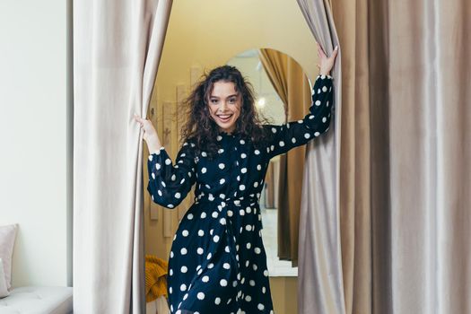 Young beautiful happy woman girl trying on a black dress with white dots near the mirror in the dressing room in a clothing store in the mall. Shows, looks at the camera, poses, brags, wants to buy