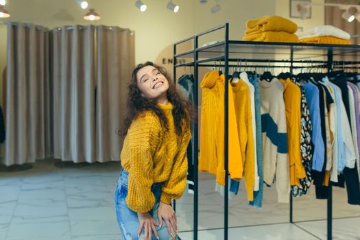 Portrait of a young beautiful curly woman, posing, dressed and posing in new clothes in a yellow warm sweater and a denim skirt in a clothing store against the background of racks with clothes on accessories, showing, bragging, looking at the camera, smiling
