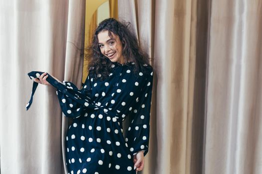 Young beautiful happy woman girl trying on a black dress with white dots near the mirror in the dressing room in a clothing store in the mall. Shows, looks at the camera, poses, brags, wants to buy