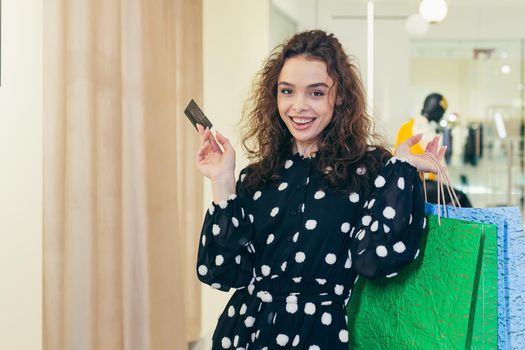 Shopping. Close-up photo. Portrait of a young beautiful curly woman holding a credit card and paper colored bags with clothes, shopping, gifts in a store in a shopping center. She looks at the camera, satisfied, happy.