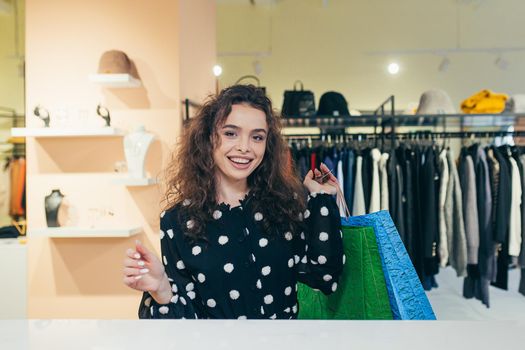 Shopaholic. Beautiful young curly woman holding colored paper bags with purchases, goods, gifts. Shows, brags, rejoices, looks at the camera in the store in the mall