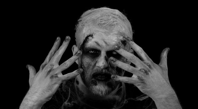 Creepy man with bloody scars face, Halloween stylish zombie make-up. Scary wounded undead guy making faces, looks at camera hiding through hands smiles terribly. Thematic party. Sinister beast monster