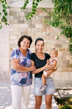 Grandmother stands next to mother holding a little girl in her arms near a stone wall. High quality photo