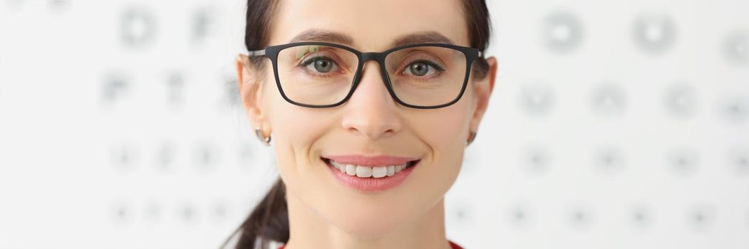 Portrait of smiling female eye doctor with eye test chart on background, female optician in glasses, appointment in eye clinic. Ophthalmologist concept