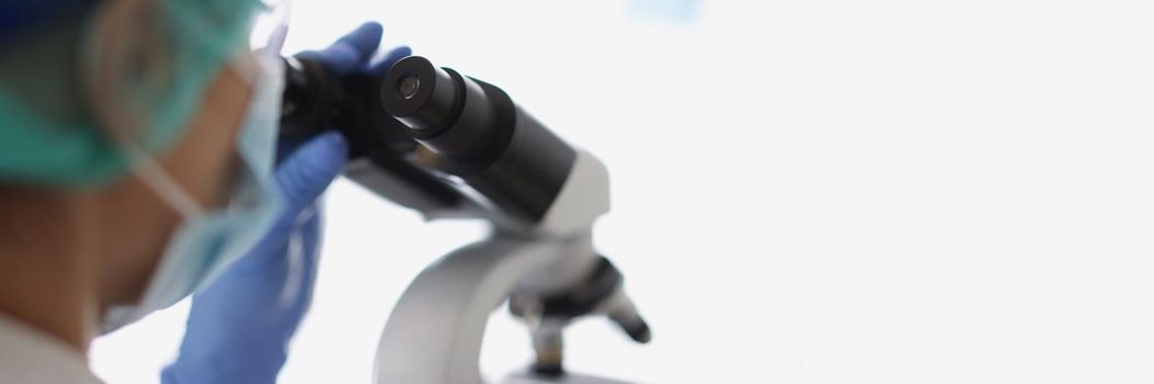 Close-up of woman chemist in blue protective glove look at microscope in chemistry lab. Scientist looking at optical microscopy metal lens. Testing concept